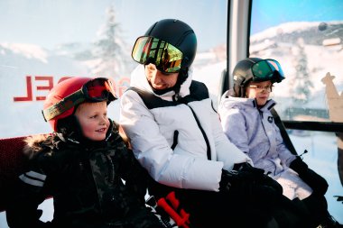 An adult and two children sitting in a gondola on a ski lift, wearing ski helmets and goggles, with snowy mountains in the background. | © Kleinwalsertal Tourismus eGen • Photographer: Oliver Farys