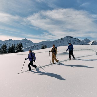 Drei Skitourengeher in schneebedeckter Berglandschaft mit Gipfeln und bewölktem Himmel, bei winterlicher Tour im Kleinwalsertal. | © Kleinwalsertal Tourismus eGen • Fotograf: Oliver Farys