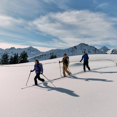 Three ski tourers in snow-covered mountain landscape with peaks and cloudy sky during winter tour in Kleinwalsertal. | © Kleinwalsertal Tourismus eGen • Photographer: Oliver Farys