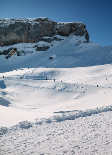 Two people walking with a dog in snowy mountains in Kleinwalsertal on a clear winter day. | © Kleinwalsertal Tourismus eGen • Photographer: Oliver Farys