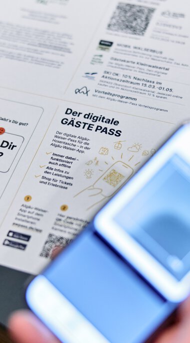Person holds a digital guest pass and reads a printed document at a table, while someone takes a photo of it with a smartphone. | © Kleinwalsertal Tourismus eGen • Photographer: Oliver Farys