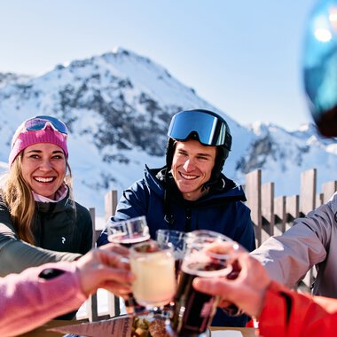 Gruppe von Freunden in Skihütte, die bei Sonnenschein mit Getränken anstoßen, Schneeberge im Hintergrund. | © Kleinwalsertal Tourismus eGen • Fotograf: Oliver Farys