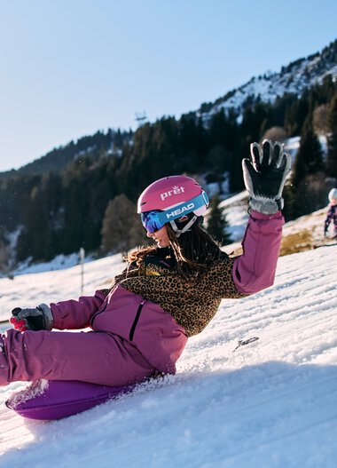 A girl sliding down a snowy slope, smiling and waving, in Kleinwalsertal with mountains in the background, enjoying winter snow tubing with other children around. | © Kleinwalsertal Tourismus eGen • Photographer: Oliver Farys