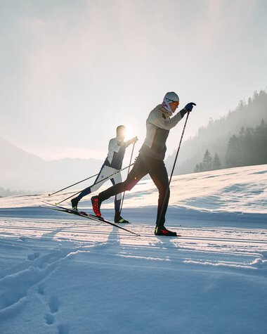 Two cross-country skiers in a snowy landscape with the sun in the background, on a trail in Kleinwalsertal. | © Kleinwalsertal Tourismus eGen • Photographer: Oliver Farys