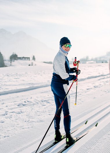 Cross-country skier on a snowy trail with mountains and winter landscape in the background, talking to someone in the foreground holding ski poles. | © Kleinwalsertal Tourismus eGen • Photographer: Oliver Farys