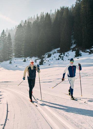 Zwei Skilangläufer in winterlicher Landschaft beim Langlaufen auf der Schwarzwasserloipe im Kleinwalsertal, umgeben von Tannen und verschneiten Bergen. | ©  Kleinwalsertal Tourismus eGen • Fotograf: Oliver Farys