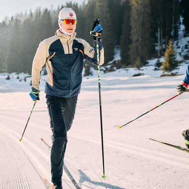Skifahrer beim Langlaufen auf der Schwarzwasserloipe im Kleinwalsertal, umgeben von verschneiten Bergen und Tannenwäldern. | © Kleinwalsertal Tourismus eGen • Fotograf: Oliver Farys