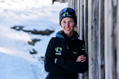 Woman in winter clothing with sunglasses and a beanie stands in the snow next to a wooden wall. | © Kleinwalsertal Tourismus eGen • Photographer: Oliver Farys