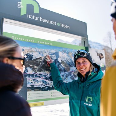 Woman in winter clothing points at a large information board with mountain images while two other people listen attentively, in snowy Kleinwalsertal. | © Kleinwalsertal Tourismus eGen • Photographer: Oliver Farys