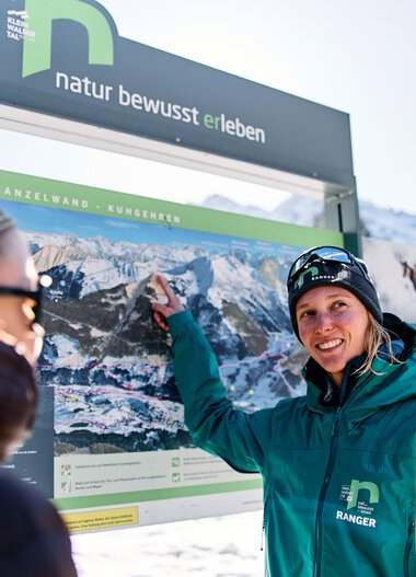 Woman in winter clothing points at a large information board with mountain images while two other people listen attentively, in snowy Kleinwalsertal. | © Kleinwalsertal Tourismus eGen • Photographer: Oliver Farys