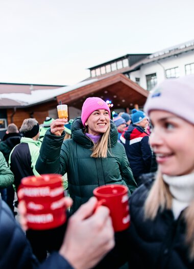 People celebrating outdoors in winter, one holding a glass, all wearing warm clothing and hats. | © Kleinwalsertal Tourismus eGen • Photographer: Oliver Farys