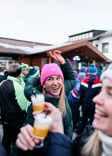 Happy woman wearing a pink beanie at snowman gathering in Kleinwalsertal, surrounded by people at a winter event. | © Kleinwalsertal Tourismus eGen • Photographer: Oliver Farys