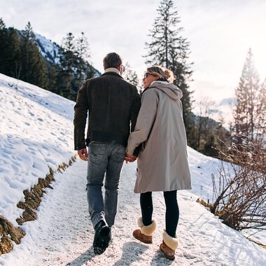 Ein Paar wandert im Schnee in einer Winterlandschaft, umgeben von Bäumen und Bergketten, bei Sonnenschein im Kleinwalsertal. | © Kleinwalsertal Tourismus eGen • Fotograf: Oliver Farys