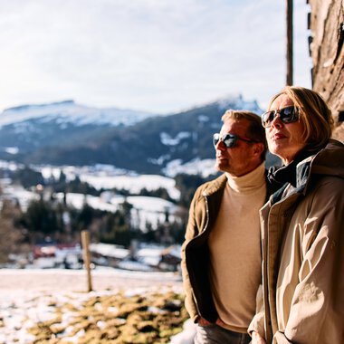 Two people stand in winter in the mountains, leaning against a wooden wall and wearing sunglasses. Snow-covered mountains are visible in the background. | © Kleinwalsertal Tourismus eGen • Photographer: Oliver Farys