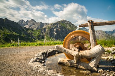 Landscape with mountains and an old water wheel amidst a green meadow under a cloudy sky. | © Kleinwalsertal Tourismus eGen • Photographer: Robert Niederwolfsgruber