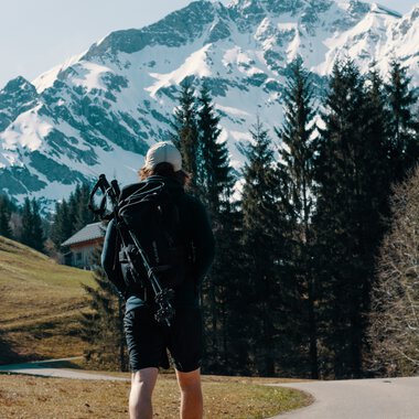 Ein Wanderer mit Rucksack und Wanderstick steht in einer Alpenlandschaft mit verschneiten Gipfeln und Tannen im Hintergrund. | © Kleinwalsertal Tourismus eGen • Fotograf: Distillery