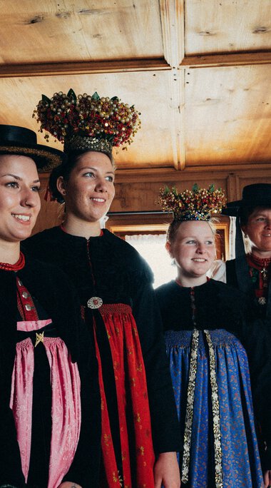 Six women in traditional Bavarian costumes standing side by side in a wooden room, smiling and looking in one direction. | ©  Kleinwalsertal Tourismus eGen • Photographer: Stefan Klauser
