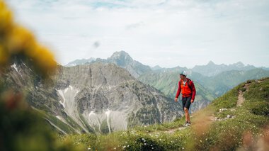 A hiker with a backpack and hat walking in the mountains, surrounded by green meadows and high peaks, with a cloudy sky in the background. | © Kleinwalsertal Tourismus eGen • Photographer: Stefan Klauser
