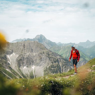 A hiker with a backpack and hat walking in the mountains, surrounded by green meadows and high peaks, with a cloudy sky in the background. | © Kleinwalsertal Tourismus eGen • Photographer: Stefan Klauser