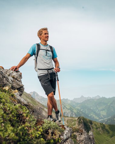 Man hiking on a rocky ledge in the mountains, carrying a backpack and hiking poles, surrounded by green hills and expansive mountain scenery. | © Kleinwalsertal Tourismus eGen • Photographer: Stefan Klauser