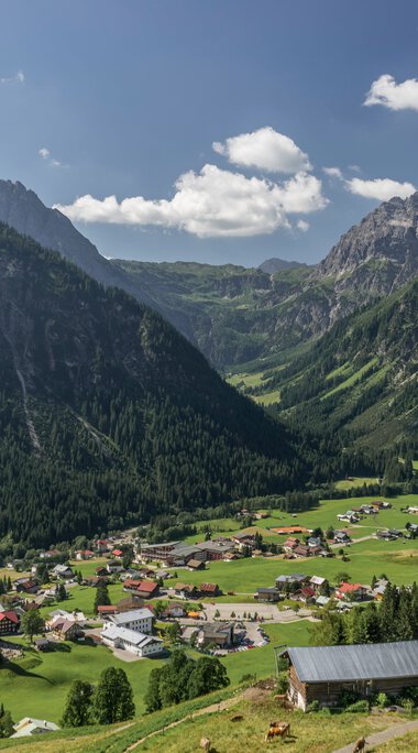Panoramaaufnahme des Bergdorfes Mittelberg in den Alpen mit grünen Hügeln, Bergen im Hintergrund und einer blauen wolkenverhangenen Himmel. | © Kleinwalsertal Tourismus eGen • Fotograf: Steffen Berschin 