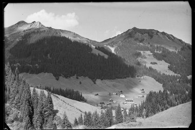 Schwarz-Weiß-Foto der Berglandschaft mit Hütten in einer talierten Landschaft, umgeben von Wäldern, mit hohen Bergen im Hintergrund. | © Kleinwalsertal Tourismus eGen • Karl Max Kessler Archiv