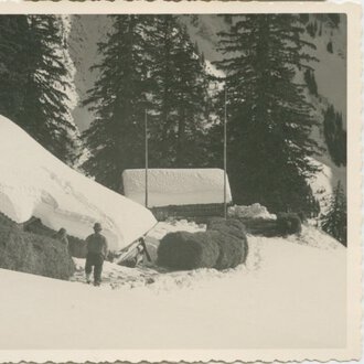 Historical black-and-white photo of a house in snowy landscape, surrounded by pine trees, with a person standing outside. | © Kleinwalsertal Tourismus eGen • Stefan Heim