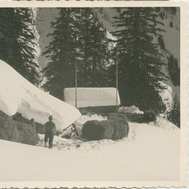 Historical black-and-white photo of a house in snowy landscape, surrounded by pine trees, with a person standing outside. | © Kleinwalsertal Tourismus eGen • Stefan Heim