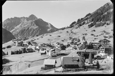 Historical black-and-white photo of a small village in the Austrian Alps with a church, surrounded by mountains and huts. | © Kleinwalsertal Tourismus eGen • Karl Max Kessler Archiv