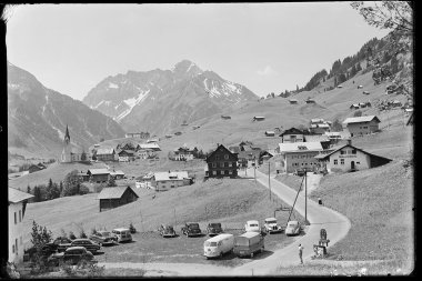 Historical black-and-white photo of the town center of Hirschegg in Kleinwalsertal, with a church, surrounding houses, cars, and the Widderstein and Hochkalber mountains in the background. | © Kleinwalsertal Tourismus eGen • Karl Max Kessler Archiv