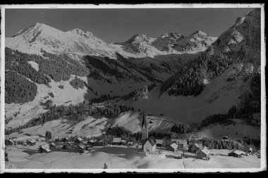 Historisches Foto des Ortzentrums von Mittelberg mit Kirche und drei Schafsalpköpfen vor verschneiten Bergen. | © Kleinwalsertal Tourismus eGen • Karl Max Kessler Archiv
