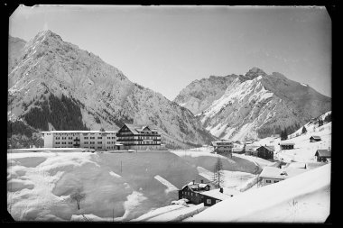 Schneebedeckte Landschaft im Mittelberg, mit Bergen im Hintergrund, einige Gebäude im Vordergrund. | © Kleinwalsertal Tourismus eGen • Karl Max Kessler Archiv