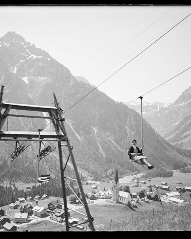 Historisches Foto eines Skiliftantriebs in einem Tal, mit Bergen im Hintergrund und kleinen Gebäuden im Vordergrund. | © Kleinwalsertal Tourismus eGen • Karl Max Kessler Archiv