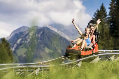 Zwei Mädchen lachen und jubeln, während sie eine Sommerrodelbahn in den Alpen hinunterfahren, umgeben von grünen Wiesen und Bergen. | © Kleinwalsertal Tourismus eGen • Oberstdorf Kleinwalsertal Bergbahnen • Fotograf: Bastian Morell