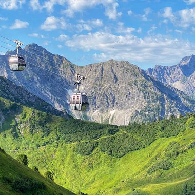 Summer Kanzelwand cable car with gondolas in the alpine landscape, green hills, mountains, and blue sky. | © Oberstdorf · Kleinwalsertal Bergbahnen