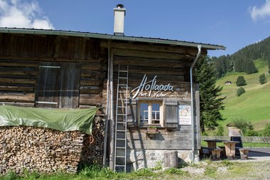 Traditional wooden hut with wood pile and ladder, surrounded by green mountains, advertising the farm shop in Kleinwalsertal. | © Kleinwalsertal Tourismus eGen • Hoflaada