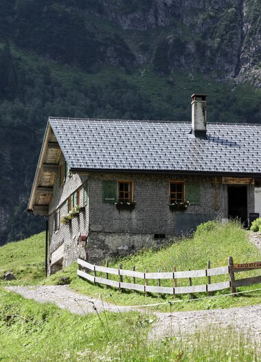Bernhards Gemstelhütte in the mountains, surrounded by green nature and forest, with terrace and umbrella. Roof made of shingles, small path leading to the hut. | © Kleinwalsertal Tourismus eGen • Bernhards Gemstelhütte • Photographer: Frank Drechsel