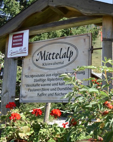 Sign for the GenussRegion Mittelalp in Kleinwalsertal, surrounded by red flowers and green plants. Wooden posts and trees in the background. | © Kleinwalsertal Tourismus eGen • Mittelalp