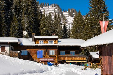 Schneebedeckte Chalets in einem winterlichen Bergdorf mit Tannenbäumen und einer steil ansteigenden Schneelandschaft im Hintergrund. | © Kleinwalsertal Tourismus eGen • Bühlalpe • Fotograf: Frank Drechsel