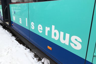 Close-up of a Walsebus in winter with snow on the ground and around the wheels, side lettering 'Walserbus' in white on a turquoise background. | © Kleinwalsertal Tourismus eGen