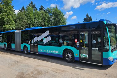 Modern water bus in blue with black and white design, parked on a street with trees and a blue sky in the background. | © Kleinwalsertal Tourismus eGen