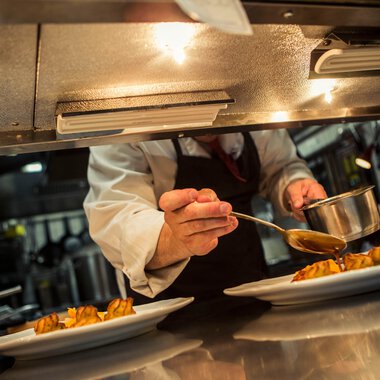 A chef carefully pouring sauce over prepared dishes in a professional kitchen. | © Kleinwalsertal Tourismus eGen • Photographer: Oliver Farys