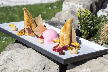 An elegant dessert on a long white plate with ice cream, fruit puree, cream, meringue, and decoration outdoors on a stone table. | © Kleinwalsertal Tourismus eGen • Photographer: Frank Drechsel