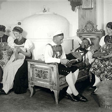 Historische Schwarz-Weiß-Aufnahme von fünf Frauen in traditionellen Trachten, die im Wohnzimmer sitzen und unterschiedliche Handarbeiten und Musikstücke machen. | © Kleinwalsertal Tourismus eGen