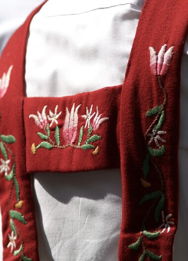 Detailaufnahme einer traditionellen Tracht mit rotem Schulterband, das reichlich bestickt ist, getragen von einer Person in Weiß. | © Kleinwalsertal Tourismus eGen • Fotograf: Hans Wiesenhofer