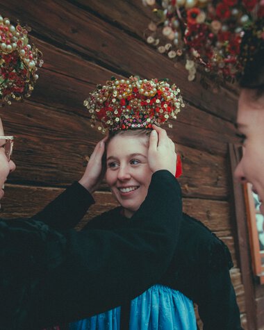 Drei Frauen in traditioneller Tracht passen einer jüngeren Frau eine festliche Kopfhaube an, im Hintergrund eine Holzvertäfelung. | © Kleinwalsertal Tourismus eGen • Fotograf: Stefan Klauser