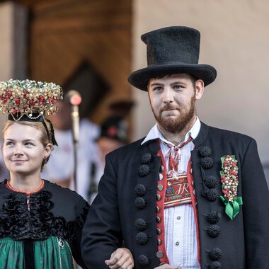 A man and a woman in traditional costumes at an alphorn festival, with the woman wearing a floral crown on her head. | © Kleinwalsertal Tourismus eGen • Photographer: Frank Drechsel