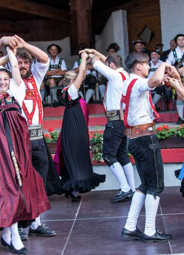 Tanzgruppe in traditionellen Trachten bei einem folkloristischen Fest im Kleinwalsertal, mit Musikern im Hintergrund. | © Kleinwalsertal Tourismus eGen • Fotograf: Frank Drechsel