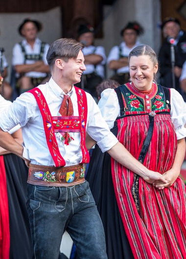 Tanzpaar in traditionellen Alpinskostümen bei einem Alphornfestival in Kleinwalsertal, alle umgeben von weiteren Tanzenden in ähnlicher Tracht. | © Kleinwalsertal Tourismus eGen • Fotograf: Frank Drechsel