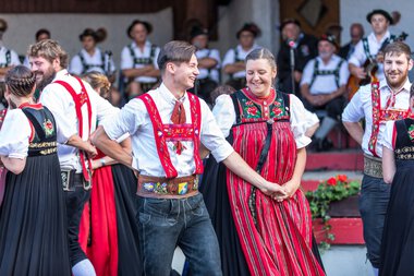 Dancing couple in traditional alpine costumes at an alphorn festival in Kleinwalsertal, surrounded by other dancers in similar attire. | © Kleinwalsertal Tourismus eGen • Photographer: Frank Drechsel